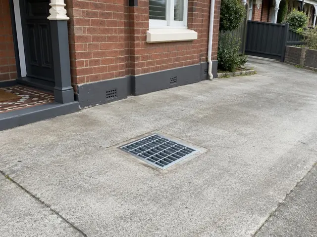 Outdoor driveway drain grate set in concrete beside a brick house and front steps in Marrickville