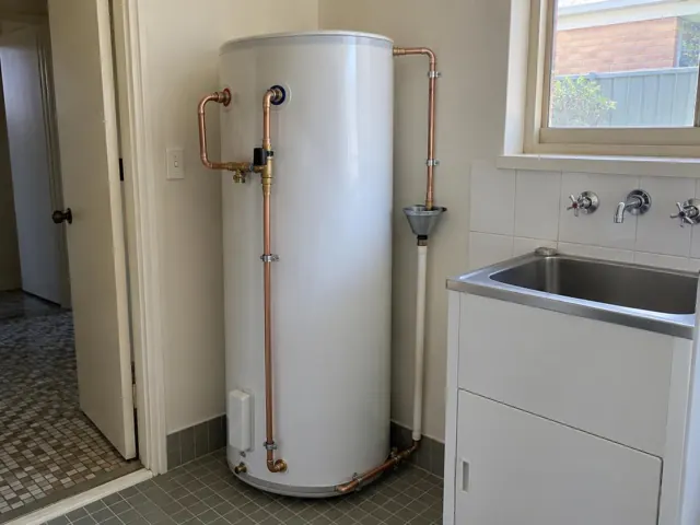 White hot water cylinder with copper pipework beside a laundry sink and tiled floor in Mascot